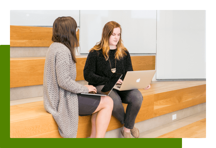two women talking on a bench together and looking at their laptops