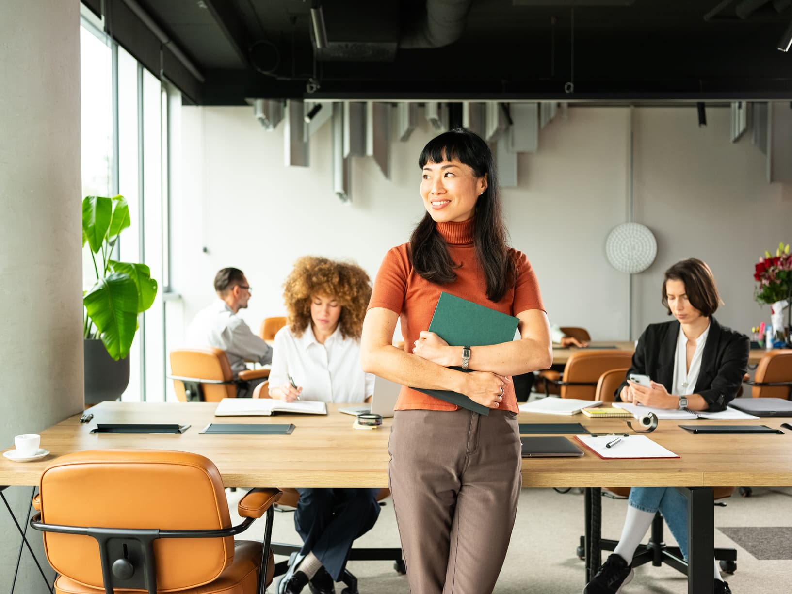 A confident woman stands smiling in an office, holding a green folder. Three colleagues work in the background at a large wooden table.